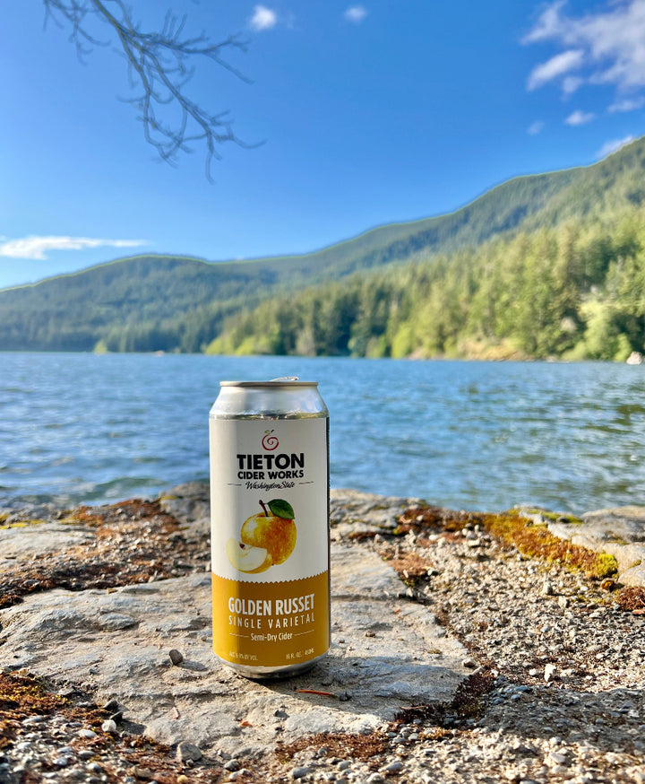 16 oz can of Tieton Cider Works Golden Russet Single Varietal Semi-Dry Cider sitting on a rock in front of a blue lake with evergreen tree covered hills in the background