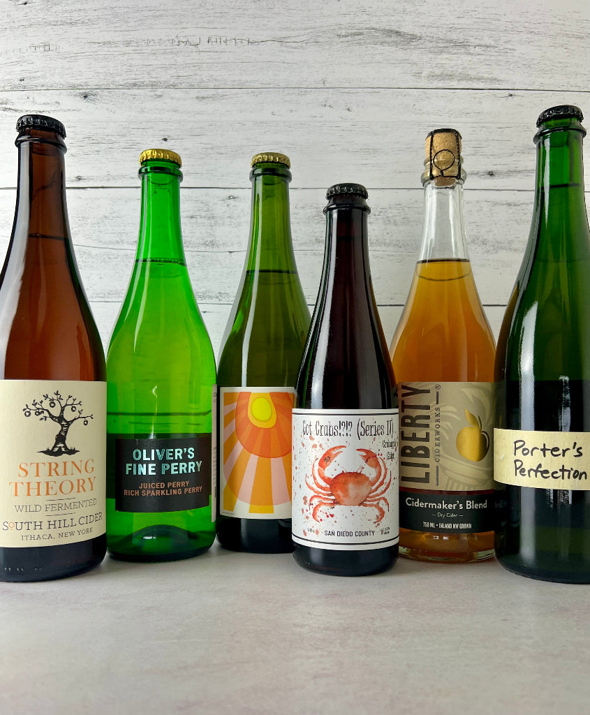 Six bottles of cider lined up against a light gray wood plank background. The bottles include String Theory, Oliver's Fine Perry, Western Cider Golden Russet, Raging Cider Got Crabs IV, Liberty Ciderworks Cidermaker's Blend, and Brooklyn Cider House Porter's Perfection.