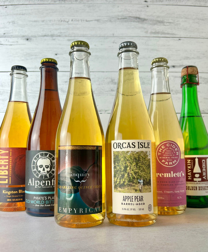 Photo of six bottles of 500 and 375 mL bottles, including Alpenfire Pirate's Plank, The Cider Farm Tremlett's, Haykin Cidery Golden Russet, Empyrical Inquiry, Orcas Isle Apple Pear Barrel-Aged cider, and Liberty Ciderworks Kingston Black