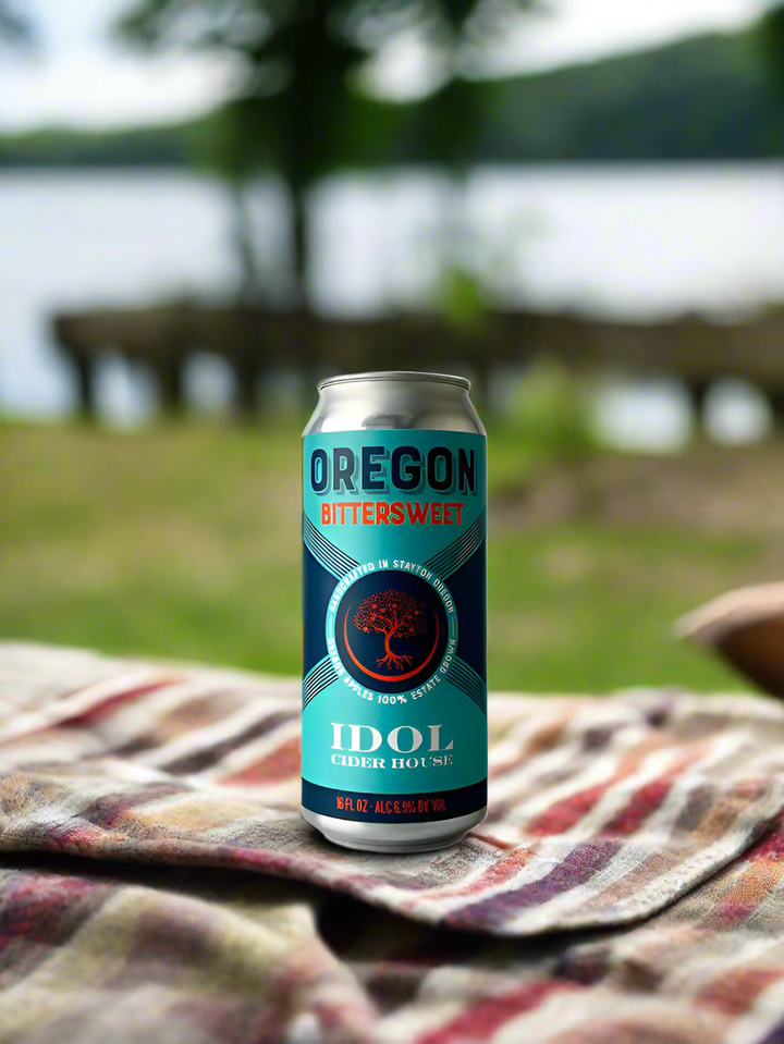 16 oz can of Idol Cider House Oregon Bittersweet cider, on a blanket in the grass with a lake in the background