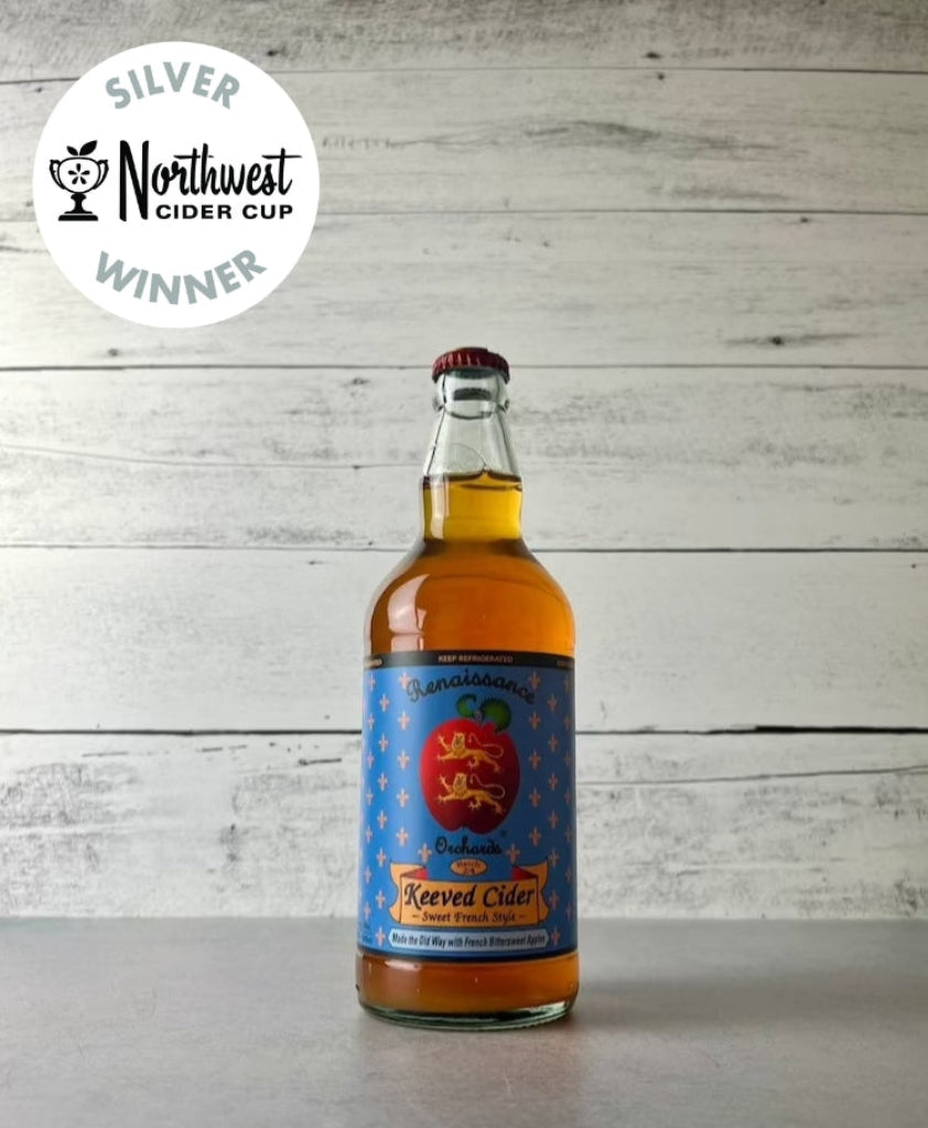 A photo of a bottle of Renaissance Orchards Keeved Cider with a label showing an apple illustration and text, placed against a wooden backdrop. There is a badge on the top left corner indicating 'Silver Northwest Cider Cup Winner'.