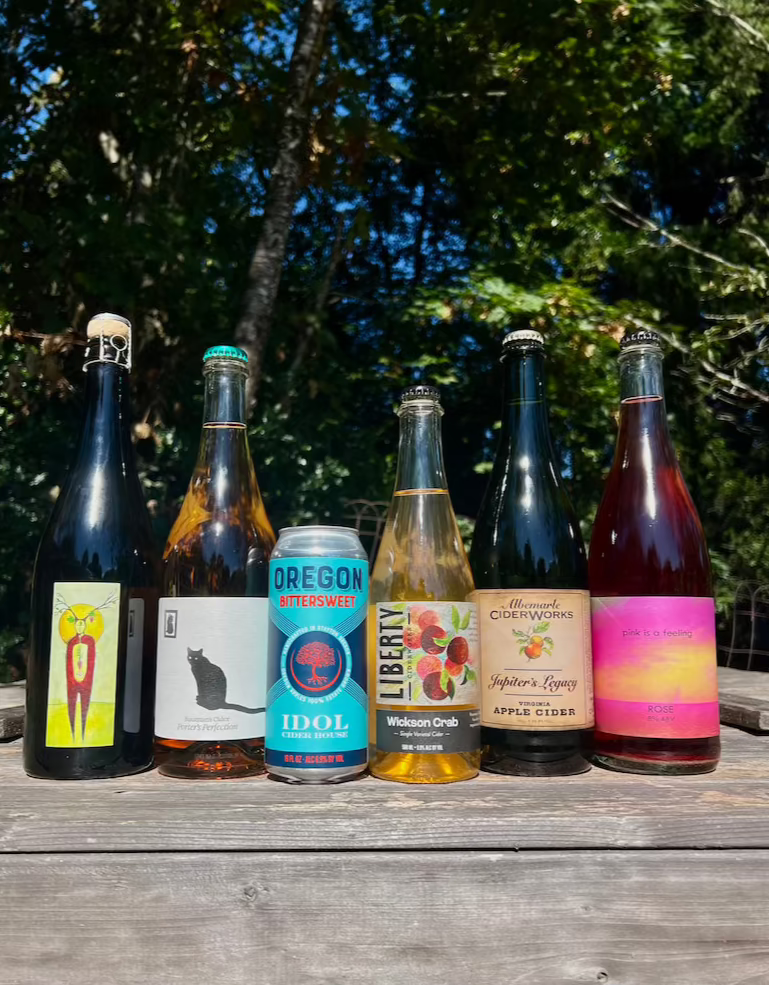 6 ciders on a wood platform with trees in the background: South Hill Cider, Bauman's Cider, Idol Ciderhouse, Liberty Ciderworks, Albemarle Cider, and Botanist & Barrel