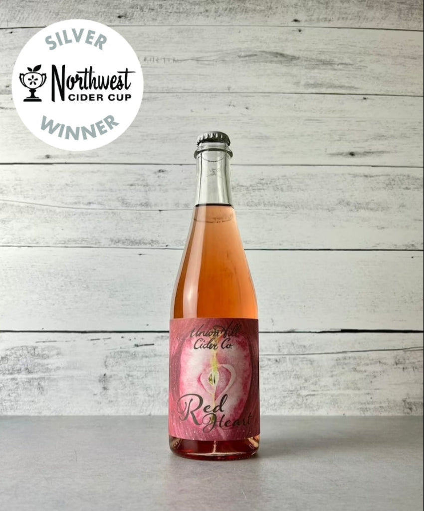Photo of a 500 mL bottle of Union Hill Cider Company's RedHeart Cider with a pink label, displaying a rosé color and indicating it is made from 100% Red Heart Apples. The bottle is presented with a Silver Northwest Cider Cup winner label.