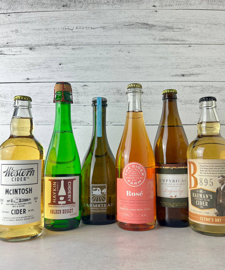 Photo of 6 500 mL bottles of cider, including Western Cider McIntosh, Haykin Cidery Golden Russet, Farmstead Cider Rafter J, The Cider Farm Rosé, Empyrical Cider Inquiry, and Bauman's Cider Clyde's Dry 
